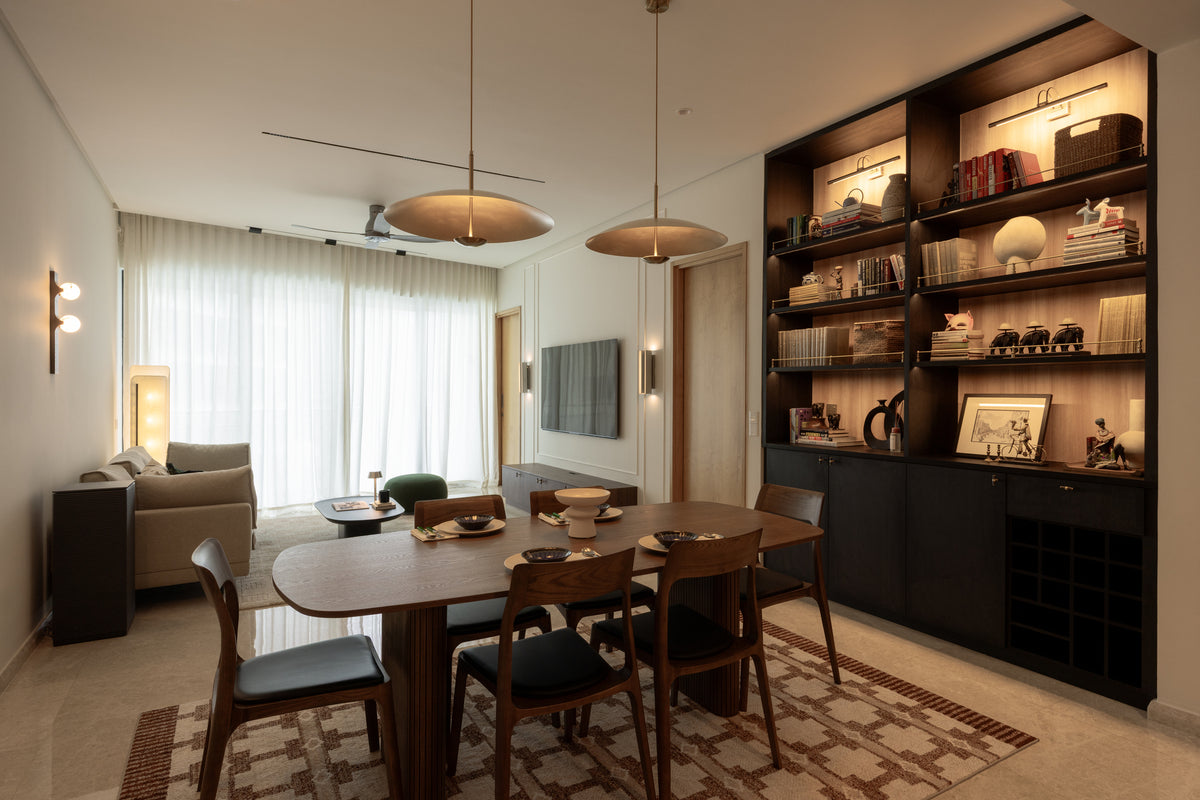 A living and dining room with a walnut table, lit dark-timber shelving, brass pendants and sheers, in a home Sundays in Bed designed at DC Residency, Damansara Heights