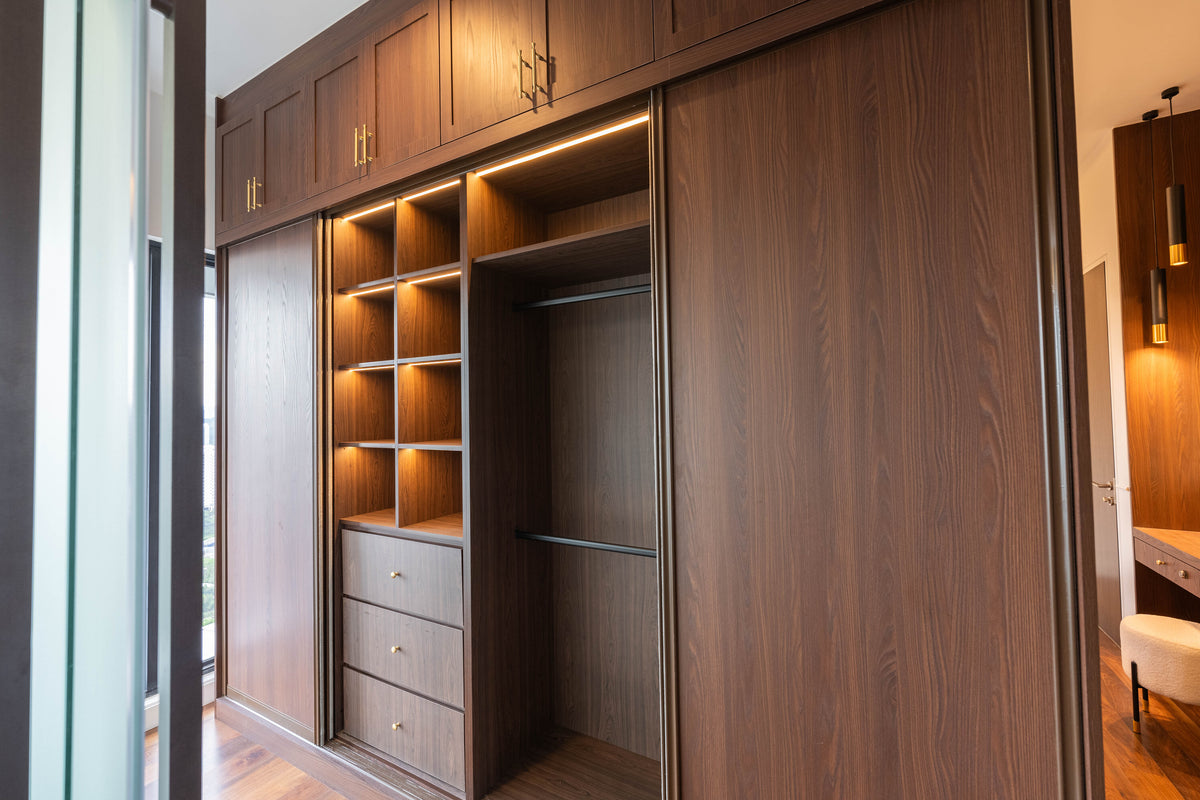Walk-in wardrobe in warm walnut with lit open shelves, a column of deep drawers, a hanging section and brass handles, in a home Sundays in Bed designed at Setia Sky Seputeh