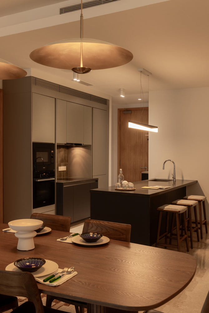 A dining table with a brass pendant, with a dark kitchen island and built-in ovens beyond, in a home Sundays in Bed designed at DC Residency, Damansara Heights