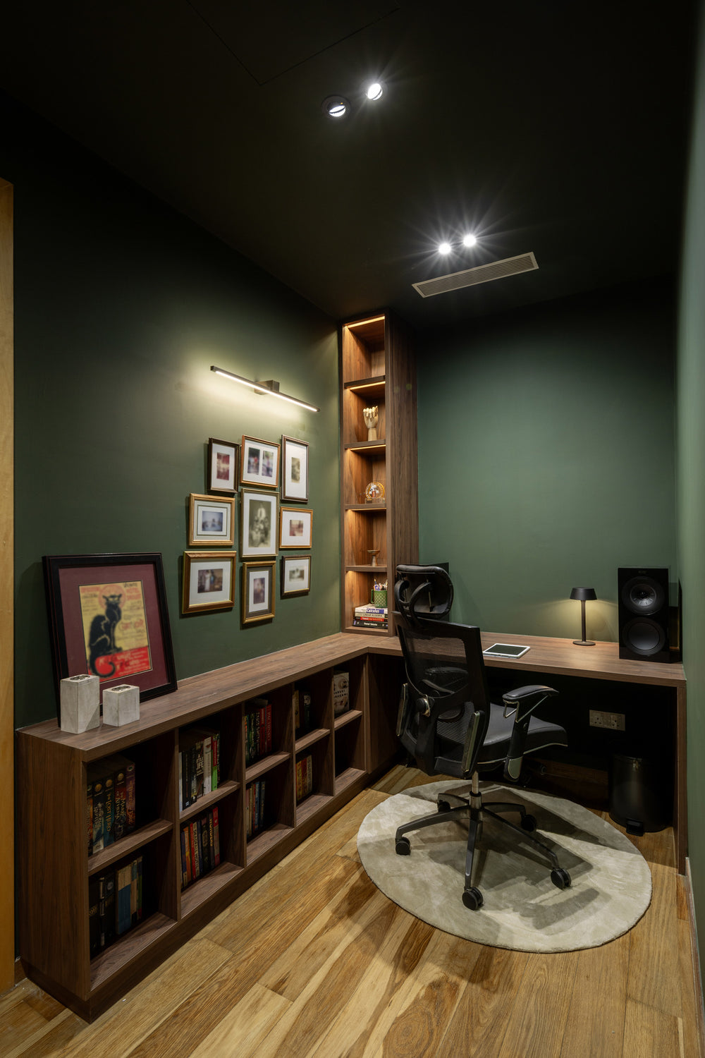 Study with deep green walls, a walnut L-shaped desk and low bookcase, framed prints and a round rug, in a home Sundays in Bed designed at DC Residensi, Damansara Heights