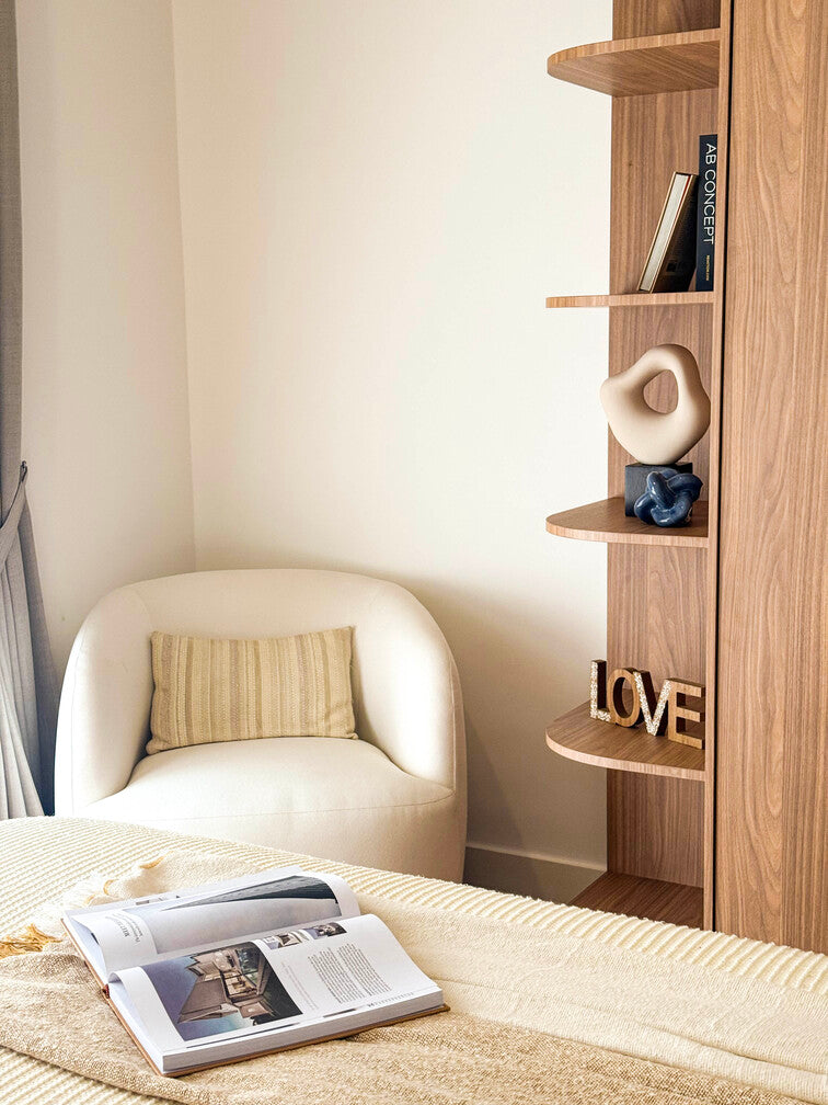 A styled living room corner: a cream armchair, layered cushions and a timber shelf with ceramics