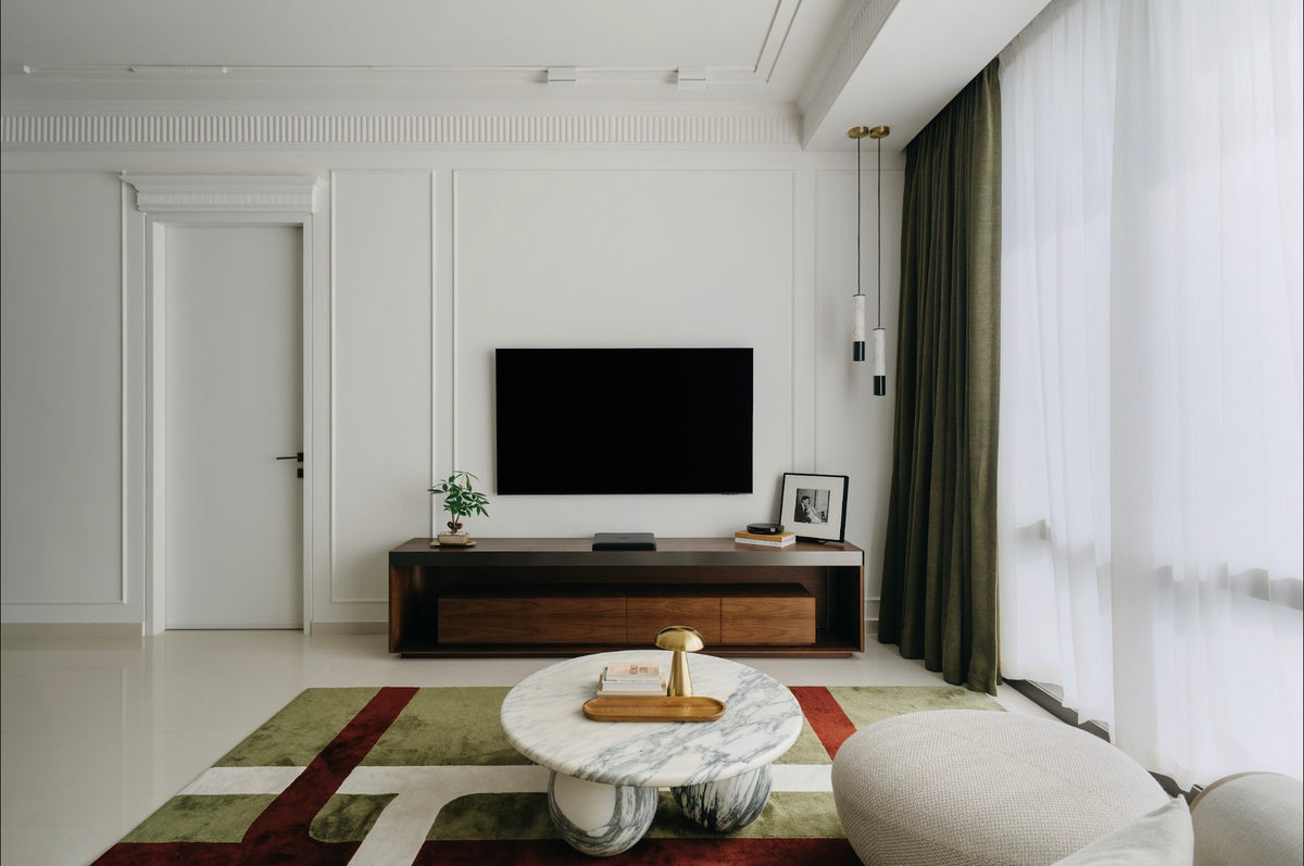 Living room with white panelled walls, sheer curtains and olive drapes along a wall of glass, a timber television console and a round stone coffee table on a green rug, designed by Sundays in Bed at Regent Suites, Damansara Heights