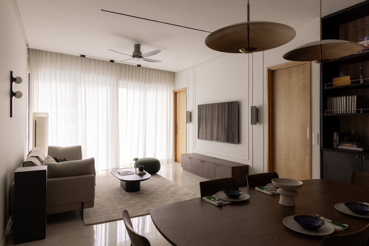 Long living and dining room seen from the dining table, with two low disc pendants, dark timber cabinetry, a sofa and sheer curtains across the far window, designed by Sundays in Bed at DC Residensi, Damansara Heights