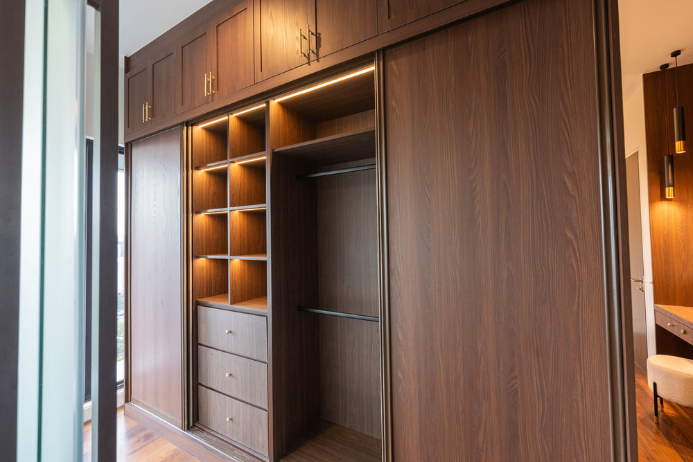 Walk-in wardrobe in warm walnut with lit open shelves, a column of deep drawers, a hanging section and brass handles, in a home Sundays in Bed designed at Setia Sky Seputeh