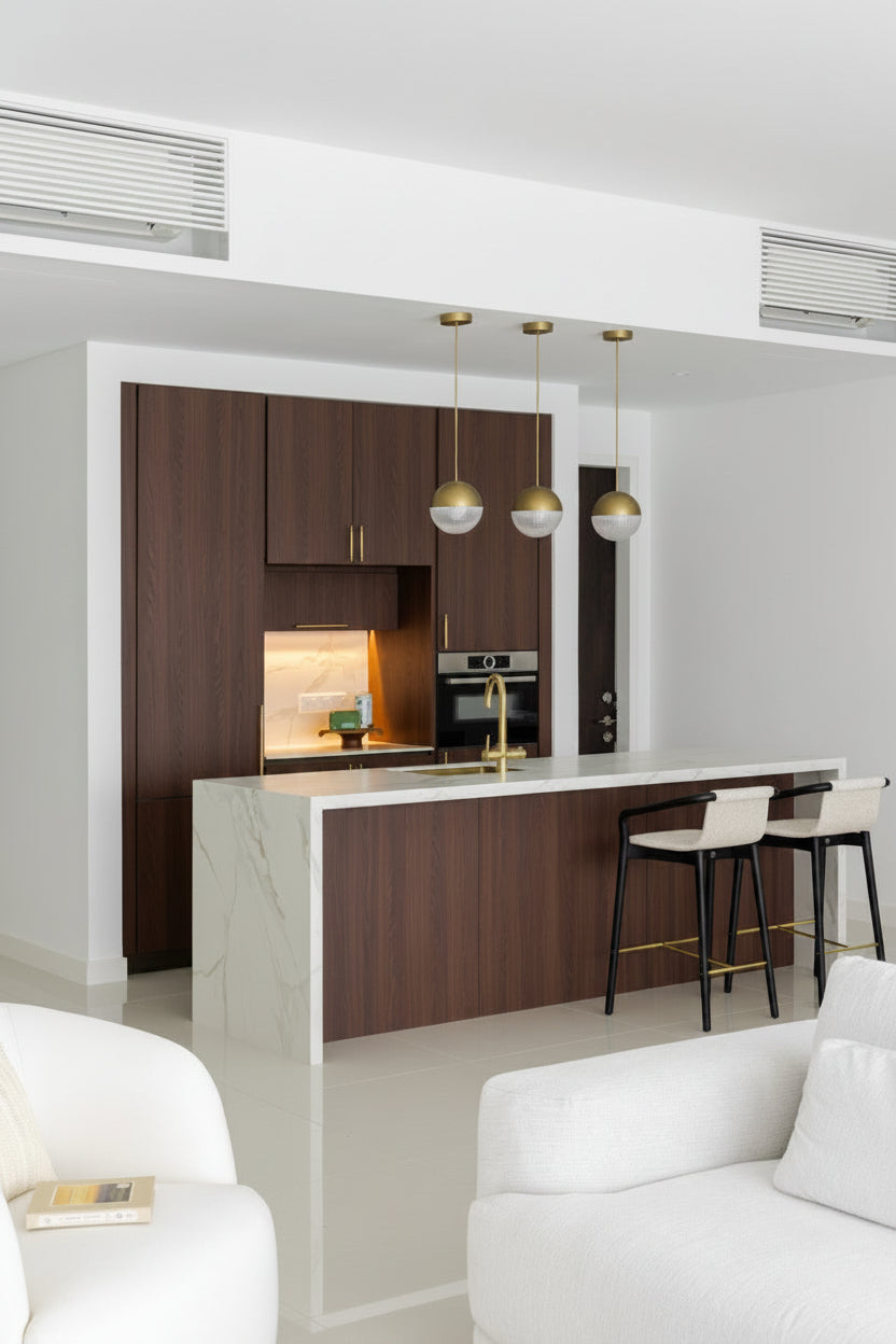 A dry kitchen with walnut cabinetry, a built-in oven, a white stone island with a brass tap and three glass pendant lights, in a home Sundays in Bed designed at Setia Sky Seputeh