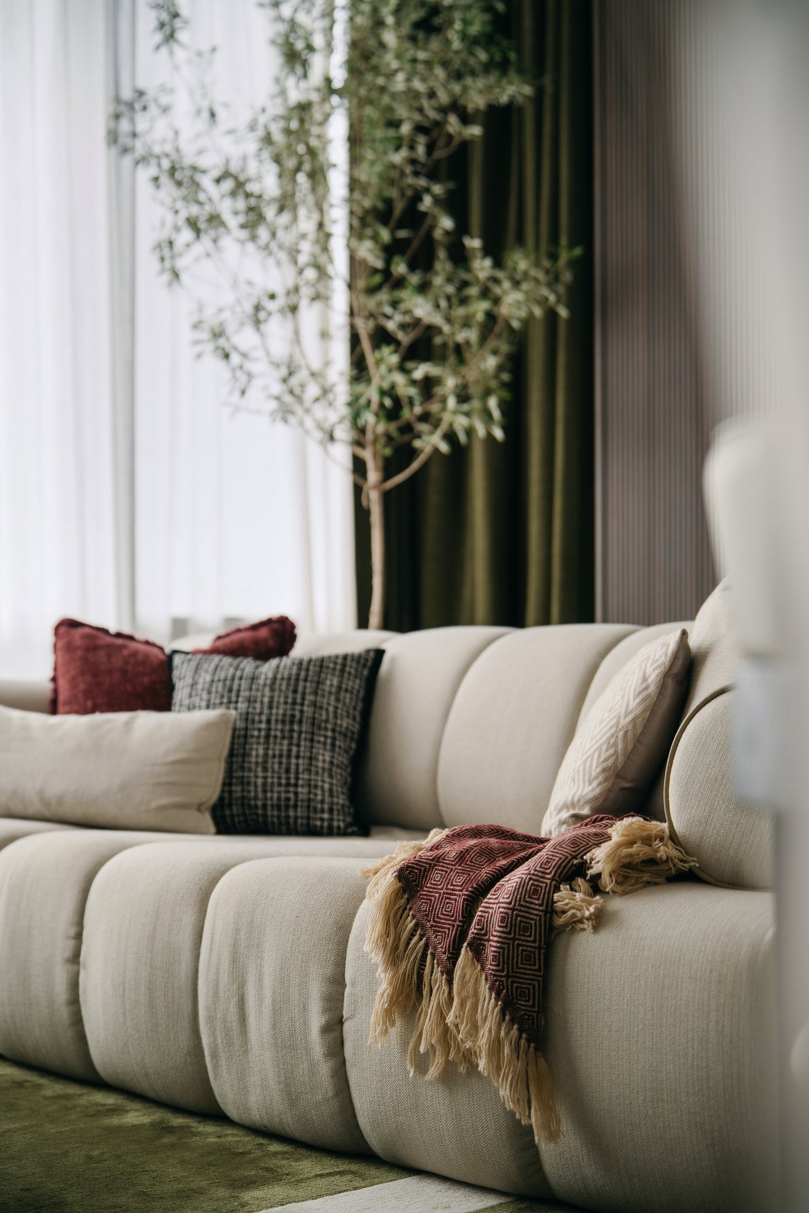 Cream channel-tufted sofa with a woven throw and cushions beside an olive tree by the window, in a living room Sundays in Bed designed at Regent Suites, Damansara Heights