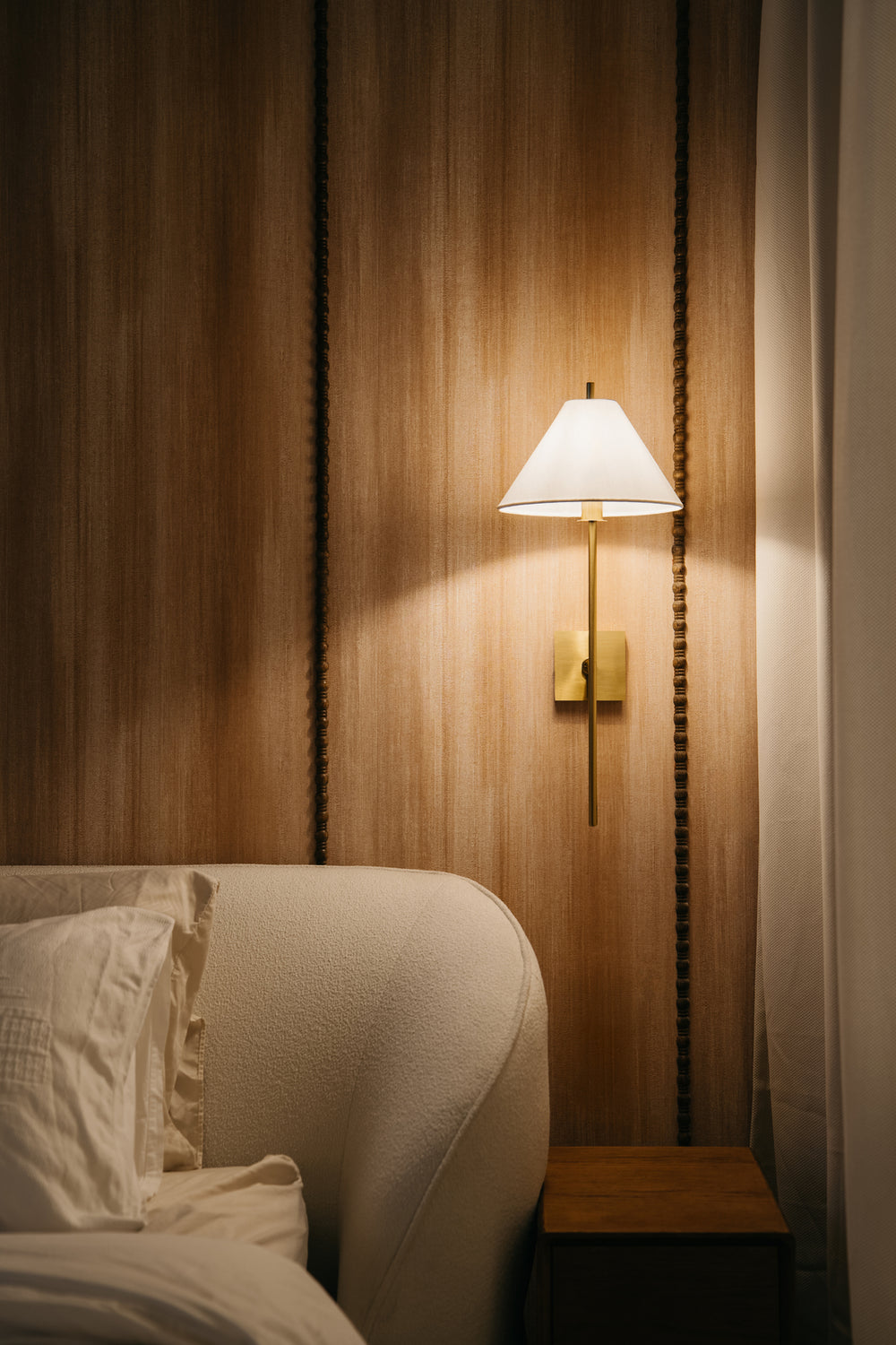Bedside corner with a brass wall light on a timber-panelled wall and a curved upholstered headboard, in a home Sundays in Bed designed at Regent Suites, Damansara Heights