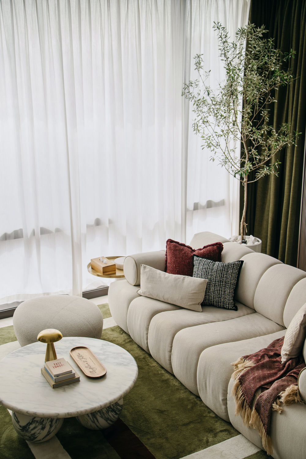 Cream channel-tufted sofa with wine and tweed cushions beside full-height sheers, olive-green curtains and an olive tree, in a living room Sundays in Bed designed at Regent Suites