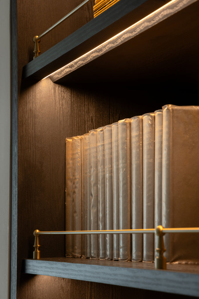 Dark timber shelving lit from beneath, with a slim brass rail across a row of pale cloth-bound books, in a home Sundays in Bed designed at DC Residensi, Damansara Heights
